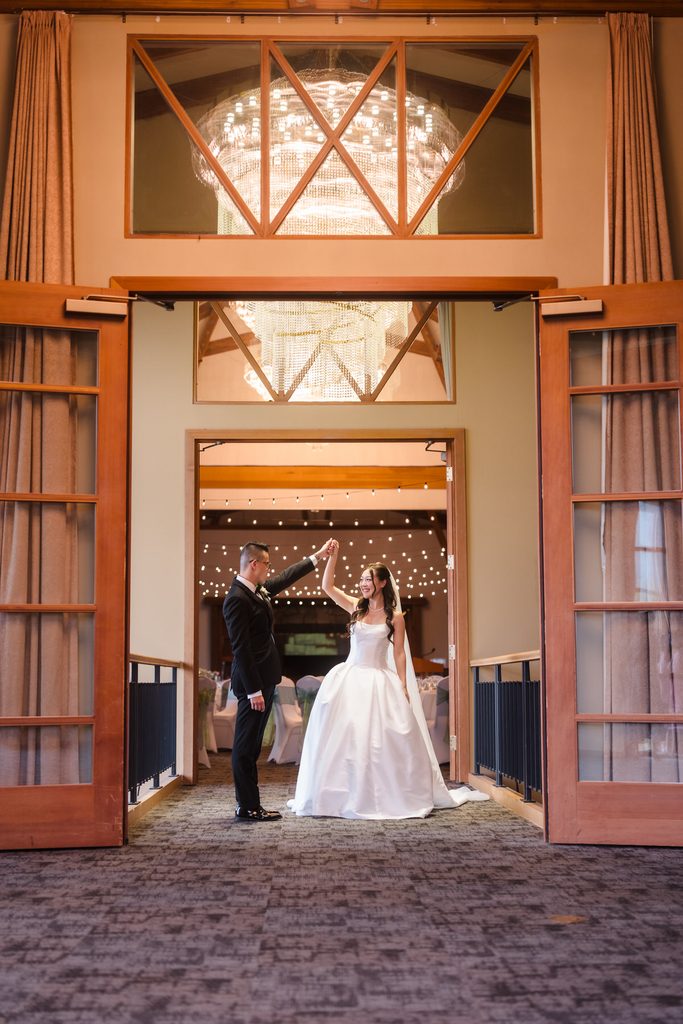 The groom raises the bride's hand as they dance in the open doorway underneath a chandelier and string lights