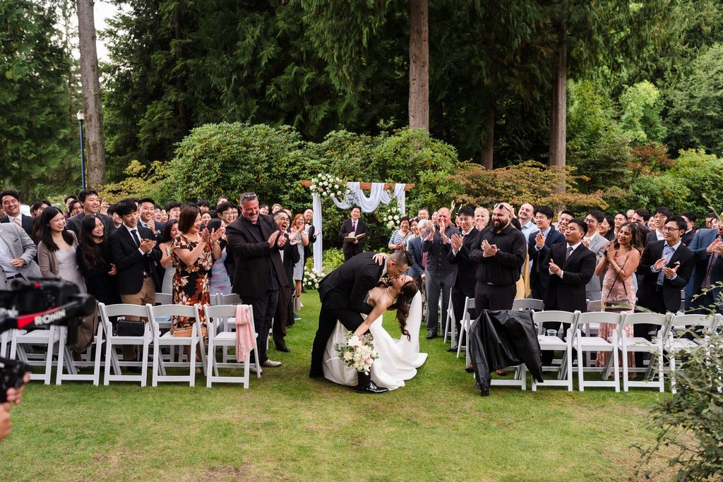 The groom dips the bride for a kiss mid aisle as guests applaud.