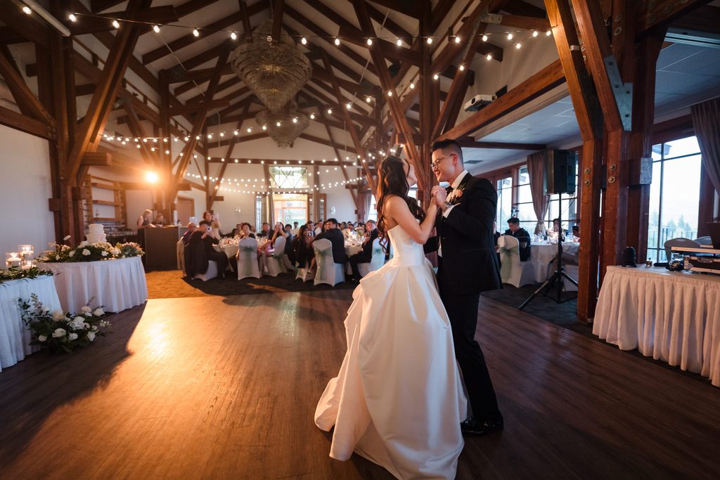 A wide shot of the timber hall shows the couples first dance near the DJ booth underneath the string lights 