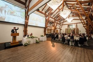 A man raising his glass to the bride and groom in a beautiful lit hall underneath the chandeliers and string lights with large vaulted ceilings.