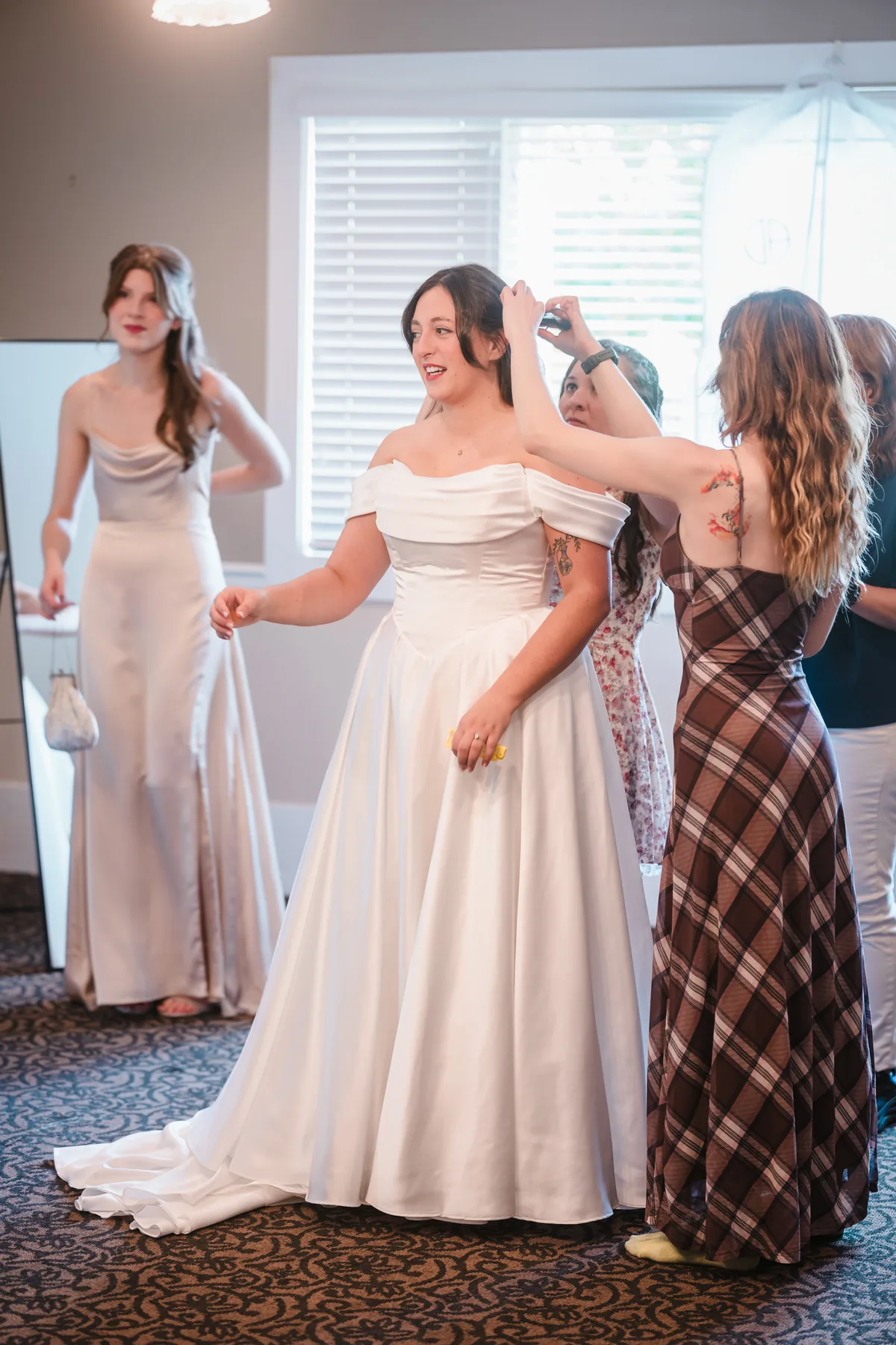 Helpers fixing the bride's hair as she stands in her gown, a bridesmaid by the mirror