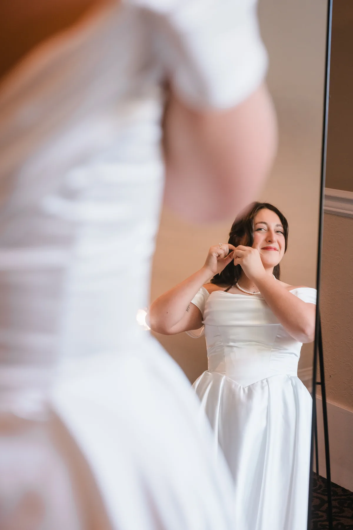 Bride's reflection in a standing mirror as she adjusts her pearl necklace, gown blurred in front