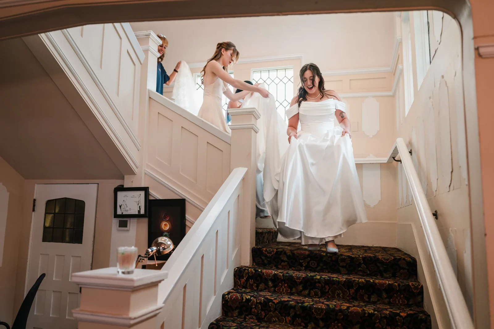 Bride smiling as she steps down carpeted stairs with a bridesmaid holding her train behind her