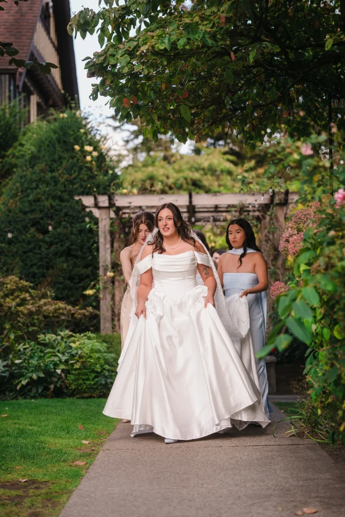 Bride holding her skirt on a garden path with bridesmaids at Hart House, Burnaby