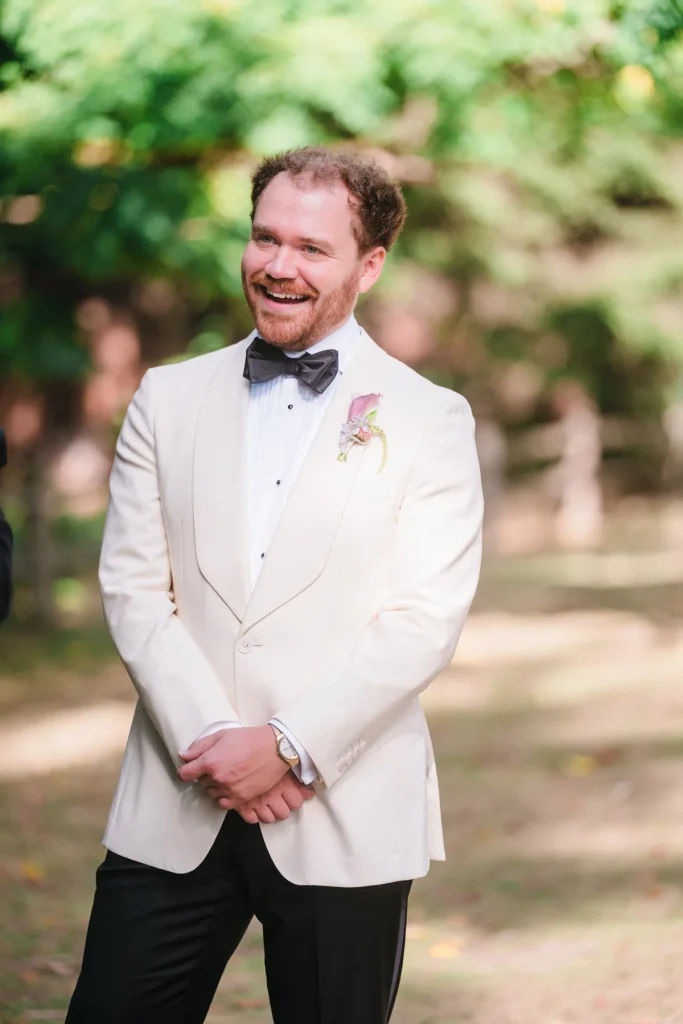 Groom laughing with a wide smile, his hands clasped, in front of blurred green trees