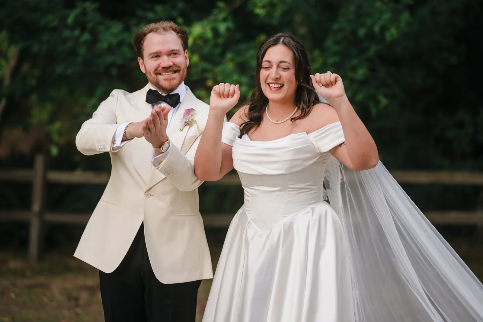 Bride with fists raised and groom clapping, both laughing in front of a wooden fence
