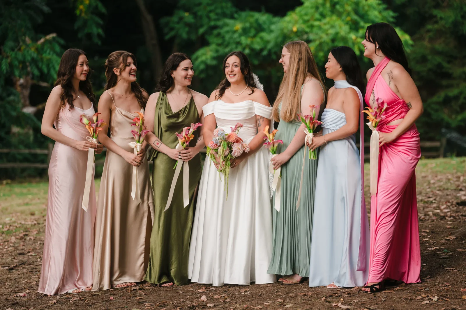 Bride and six bridesmaids laughing in a row in pink, green, blue and olive dresses