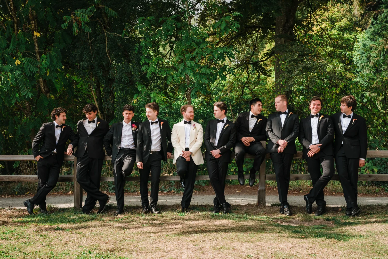 Groomsmen on a wooden fence turned toward the man in the cream jacket, smiling