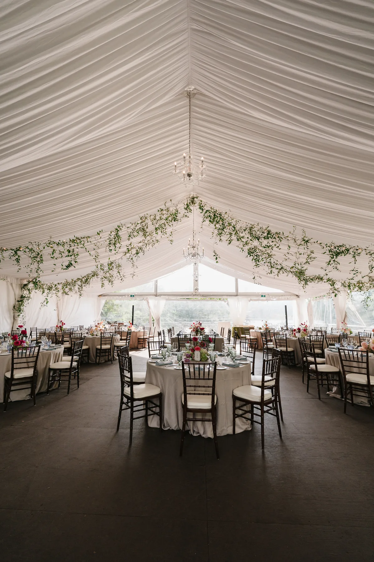 Empty reception tent with round tables, chandeliers and a leafy garland along the draped ceiling