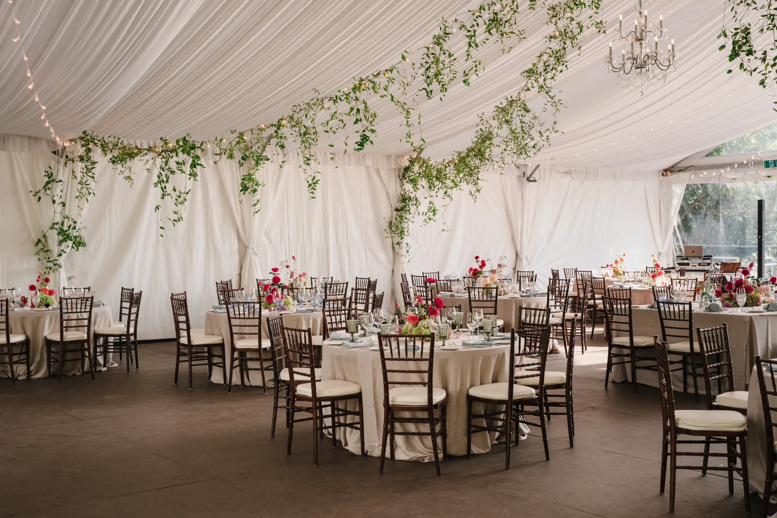 Reception tent with set tables, wooden chairs and a leafy garland hanging from the ceiling