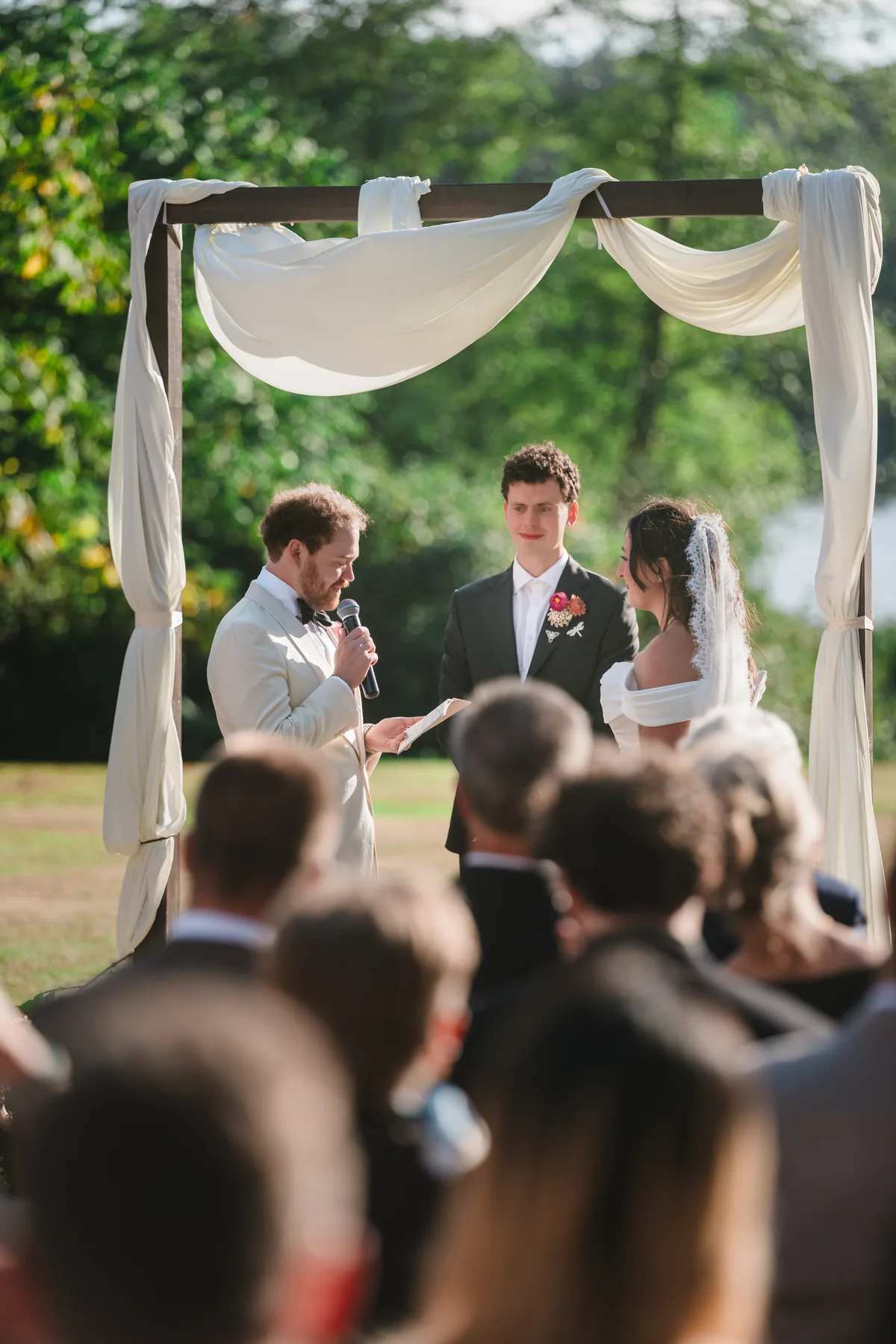 Groom reads vows at the wooden arch while guests' heads fill the foreground