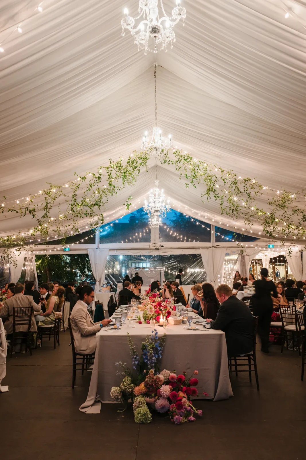 Tall view of a glowing dinner tent with chandeliers, garland and a long table of guests