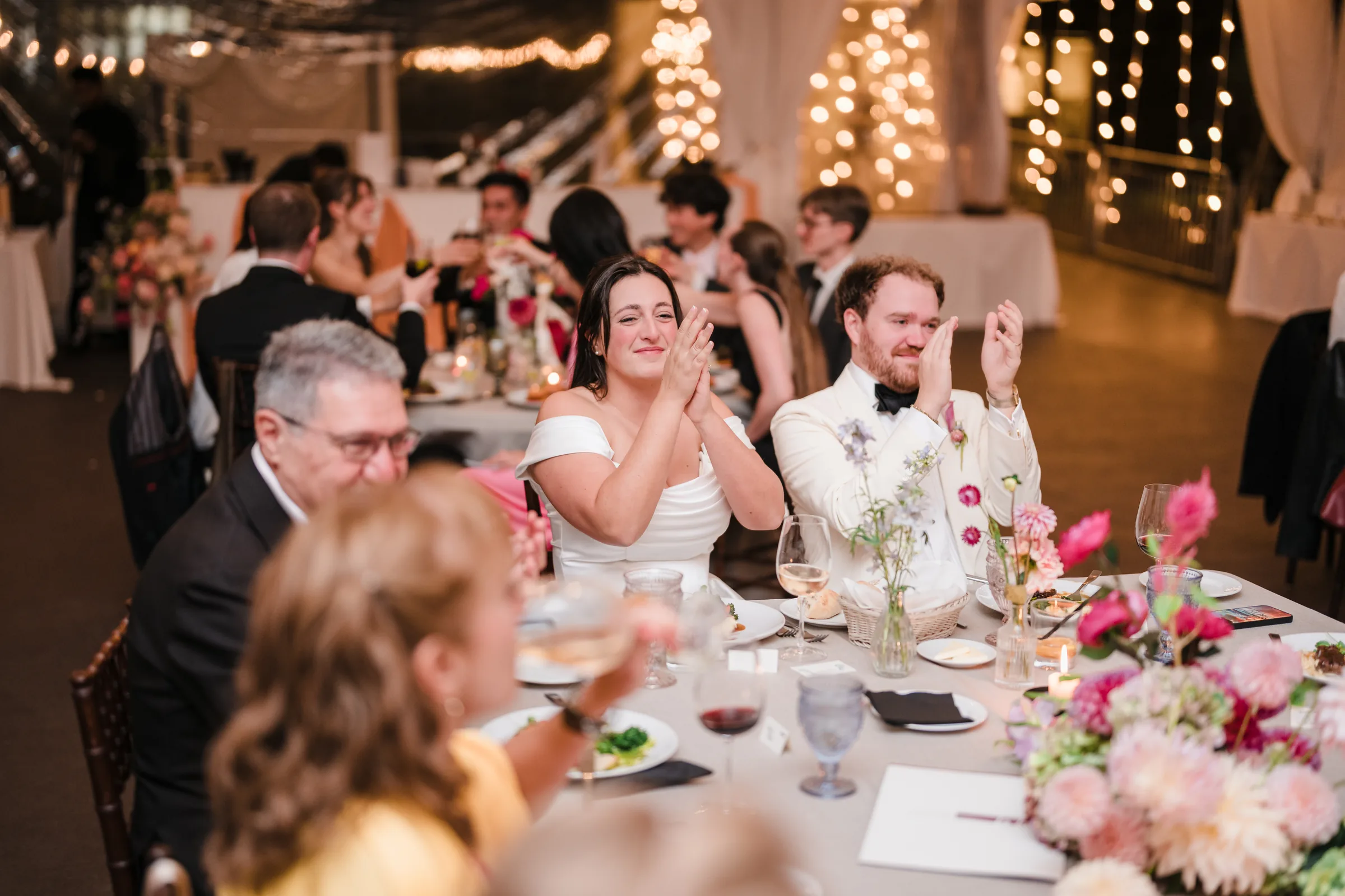 Bride and groom applauding at their table with pink and white flowers in front