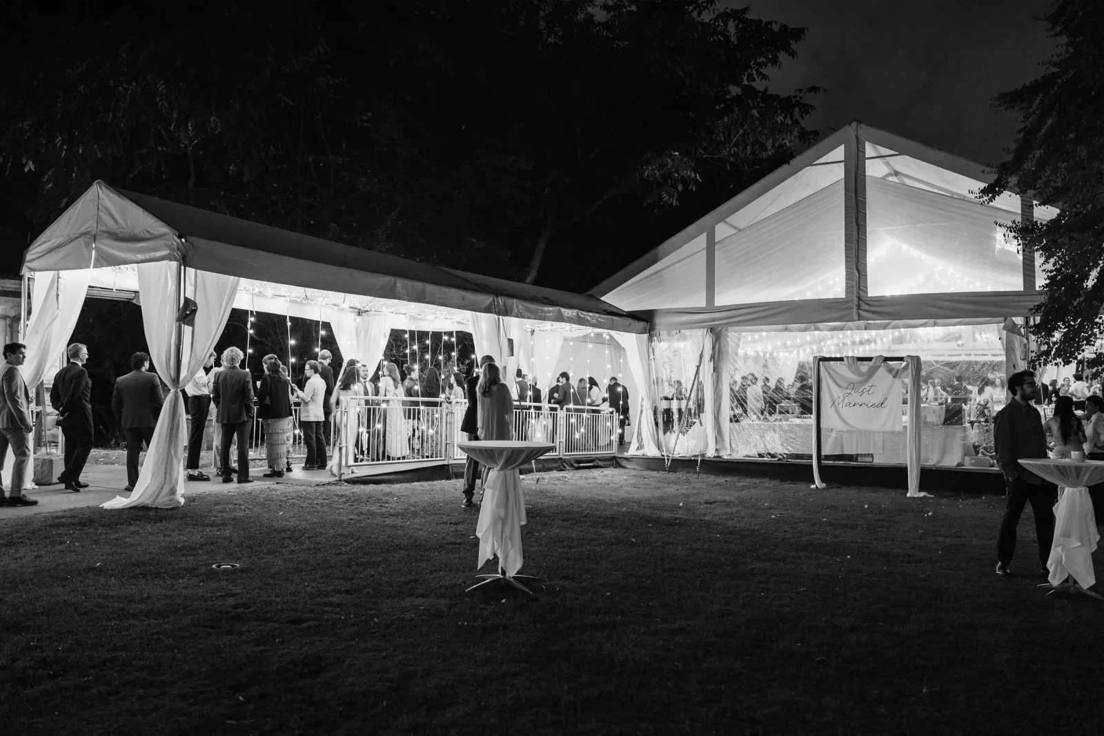 Guests mingling around a lit tent and cocktail tables at night, black and white